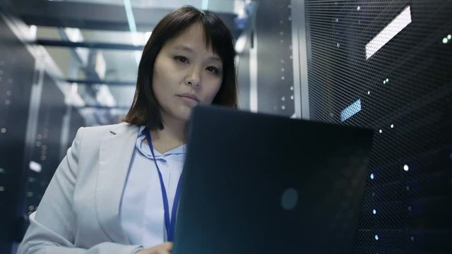 Close-up of Female Asian IT Engineer Working on a Laptop in Data Center full of Rack Servers. Shot on RED EPIC-W 8K Helium Cinema Camera.