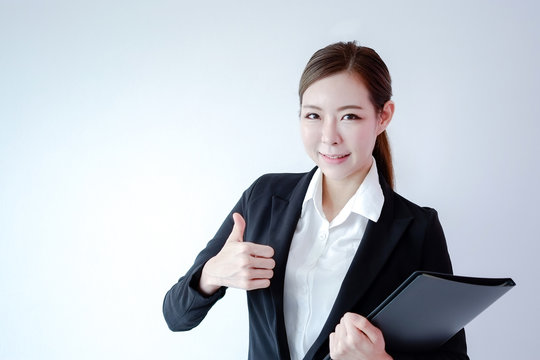 Smiling beautiful young asisn woman in business suit showing thumb up and holding document file. studio shot on white background.