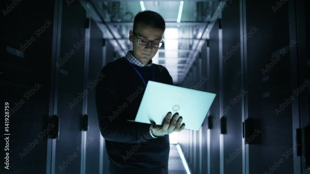 Wall mural Mid Shot of Server Engineer Reading Data Of Rack Server and Analysing it on a Laptop. He's Working in Data Center With Rows of Rack Servers.  Shot on RED EPIC-W 8K Helium Cinema Camera.