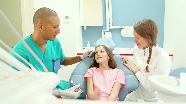 Lovely little girl talks with female dentist and male surgeon, shows her aching tooth