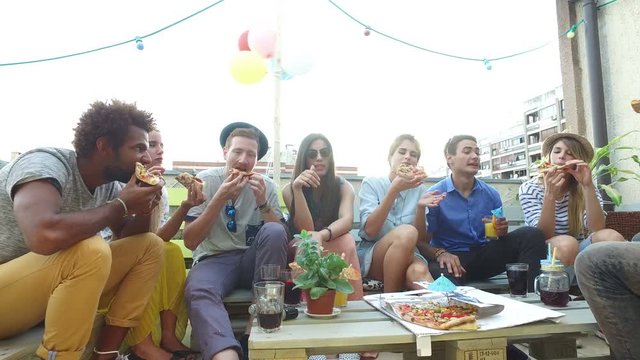 Young cheerful people sitting around the table and eating pizza on the rooftop terrace