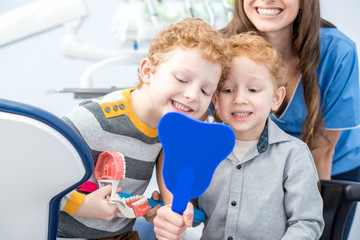 Young boys looking at the mirror with toothy smile sitting with dentist at the dental office