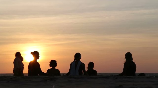 Women meditating on the beach at sunset, with people passing.