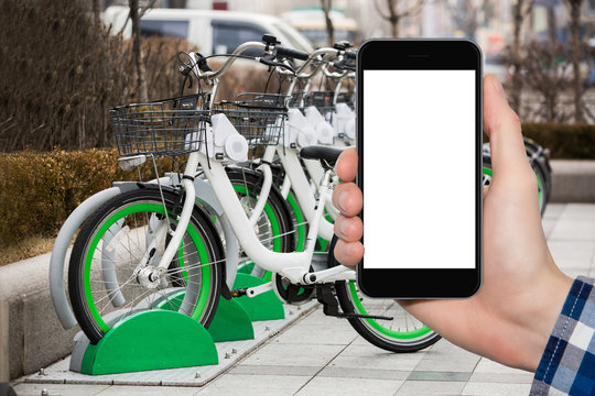 Hand With Phone On A Background Of Bicycle Rental. White Screen, You Can Insert Your Own Image Or Text Here.