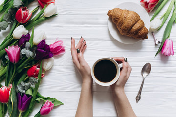 Female hands holding cup of coffee and excellent breakfast of sweet croissant on the smartphone and tulips background for Mother's Day.