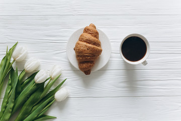 Cup of coffee with croissant, fresh tulips, smartphone and notebook sheet of paper on wooden background for Mother's Day.