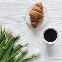 Cup of coffee with croissant, fresh tulips, smartphone and notebook sheet of paper on wooden background for Mother's Day.