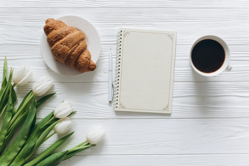 Cup of coffee with croissant, fresh tulips, smartphone and notebook sheet of paper on wooden background for Mother's Day.