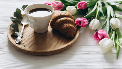 Cup of coffee with croissant and fresh tulips on wooden background for Mother's Day.