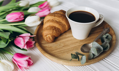 Cup of coffee with croissant and fresh tulips on wooden background for Mother's Day.