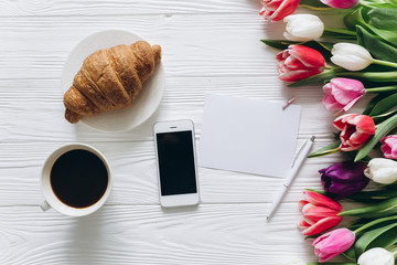 Cup of coffee with croissant, fresh tulips, smartphone and notebook sheet of paper on wooden background for Mother's Day.