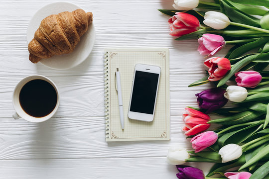 Cup of coffee with croissant, fresh tulips, smartphone and notebook sheet of paper on wooden background for Mother's Day.