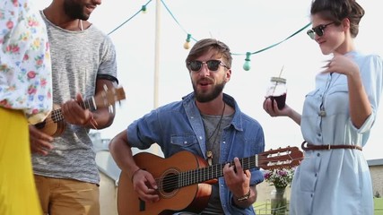 Two handsome musicians playing guitar and ukulele on the rooftop terrace on sunny summer day, friends dancing around them