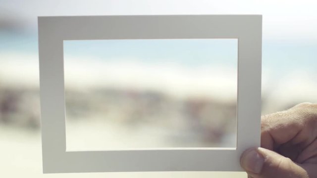 Man's hand holds empty frame for a photo against the blue sea, beach and foam waves. Closeup