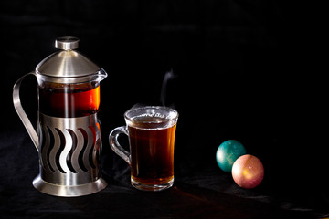 French press with tea and a Cup of tea, Easter eggs on black background.