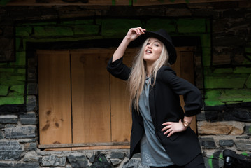 Fototapeta na wymiar Cute blond teen with long straight hair wear black jacket and hat standing against stone wall with big window