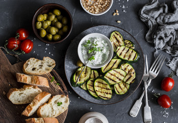 Fototapeta na wymiar Grilled zucchini, olives, tomatoes, ciabatta - simple snack or appetizer. Mediterranean style food. On a dark background, top view. Flat lay