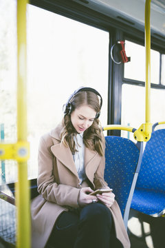 Beautiful happy young woman sitting in city bus, looking at mobile phone and listening to music. 
