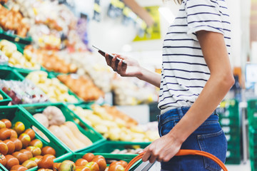 Young woman shopping healthy food in supermarket blur background. Female hands buy nature products using smart phone in store. Hipster at grocery using mobile. Person comparing price of produce