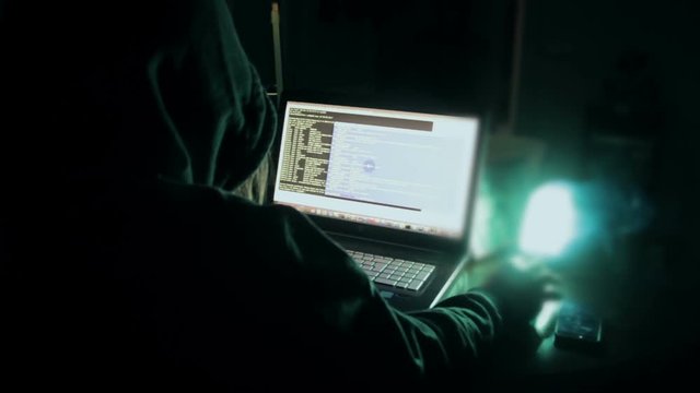 Hacker in hood cracking code using laptop and computers from his dark hacker room