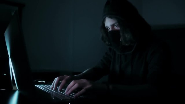 Hacker in hood cracking code using laptop and computers from his dark hacker room