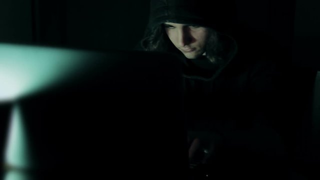 Hacker in hood cracking code using laptop and computers from his dark hacker room