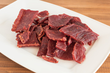 Beef jerky pieces on white dish on wooden background.