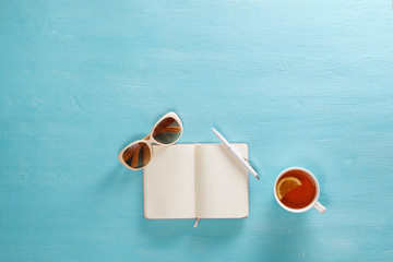 Opened notebook with pen, cup of tea and sunglasses on blue wooden table. Top view. Writing, blogging concept