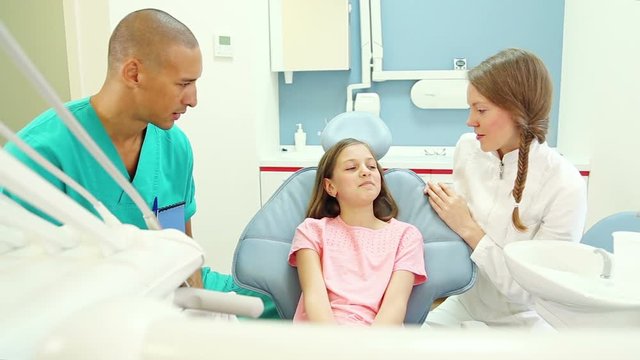 Lovely little girl talks with female dentist and male surgeon, shows her aching tooth