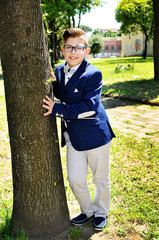 smiling boy standing near a tree at a park wearing a jacket and a bow tie shirt