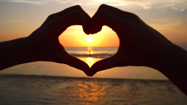 Young woman shapes heart with hands over the sunset by the ocean