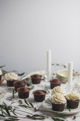 food photo, chocolate muffins and cupcakes white cream in ceramic plates of handwork on a white wooden background with green eucalyptus and baby's breath with candles top view