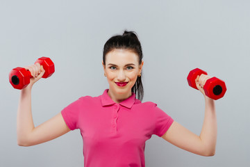 Young beautiful woman playing with two dumbbells on grey background