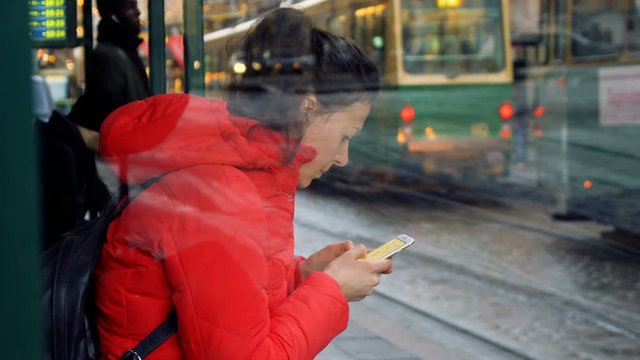 view through the glass of young travel woman in red jacket use telephone mobile for map online sitting on tram station waiting for transport in Helsinki city