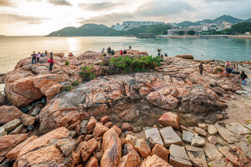 Fototapeta premium People have rest on the rocks of Stanley Bay shore in Hong Kong. Beautiful scenic sunset landscape with water, mountains, rocks and buildings
