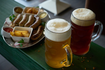 two glasses of light and dark beer with thick white foam on a wooden green the bar next to a plate of appetizers: fried potatoes and sausages and sauces
