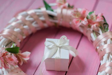 White gift box with white bow inside wreath with flowers on wooden pink background