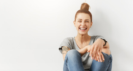 Hipster girl with hair bun wearing stylish clothing sitting on floor against white studio wall background with copy space for your advertisement while enjoying free time at home, having happy look