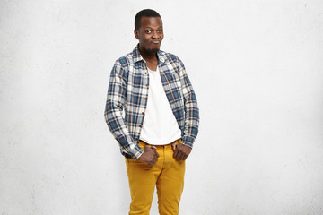 Confused frustrated dark-skinned male wearing mustard pants and checkered shirt over white t-shirt, pursuing lips and raising brows, his look and posture expressing indifference and disinterest