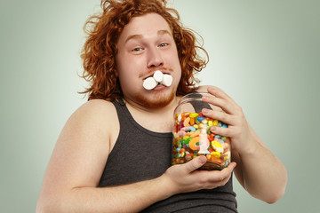 Hungry funny redhead overweight Caucasian male looking at camera with mouth full with marshmallow,...