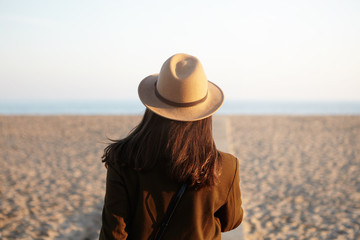 Rear view of young female with loose hair dressed in stylish warm clothing walking down coastline along boardwalk, spending holidays by ocean. Unrecognizable girl enjoying evening on city beach