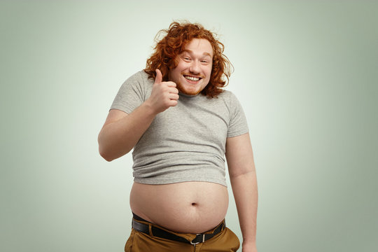 People, unhealthy lifestyle, diet and obesity concept. Fat overweight redhead Caucasian man in undersized t-shirt showing thumps up and smiling happily while his big stomach hanging out of pants