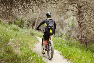 Fototapeta na wymiar Rear view of unrecognizable rider cycling on his booster bicycle on trail outdoors, enjoying fresh air in forest. Male biker riding motor-powered electric bike during morning workout in countryside