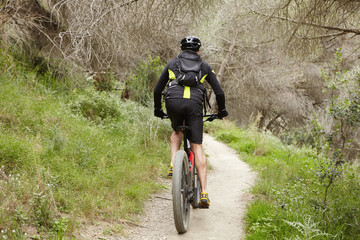 Back shot of unrecognizable cyclist riding two-wheeled electric vehicle along trail in forest alone in the morning. Stylish male biker in black sports clothes cycling on pedelec on path among trees