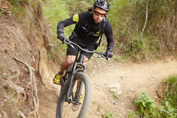 Fototapeta na wymiar Male rider wearing black cycling clothing, helmet and glasses riding along trail in forest or park, performing bike tricks on his two-wheeled electric vehicle, getting over obstacles on his way