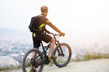 Sports, healthy lifestyle, inspiration and achievement concept. Rear view of young biker on mountain electric bicycle standing on top of hill and contemplating beautiful view of city down on sunny day