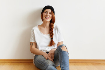 Fototapeta na wymiar Beautiful fashionable young woman with happy smile wearing stylish hat, oversize t-shirt and ragged jeans, relaxing indoors, leaning back on white wall, feeling happy and carefree on her day-off