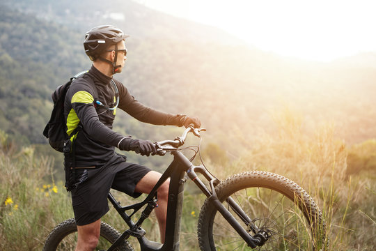 Side view of young stylish male biker in black sportswear standing on top of hill with his electric two-wheeled vehicle admiring beautiful sunset while cycling high in mountains. Flare sun effect
