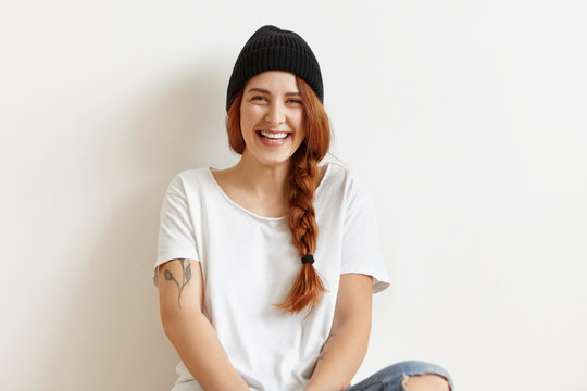 People and lifestyle concept. Indoor isolated shot of fashionable happy redhead Caucasian girl wearing black hat, white oversize t-shirt and ragged jeans having rest at home, sitting on wooden floor