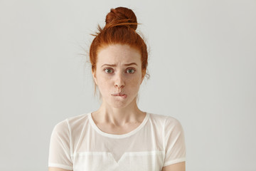 Fearful young Caucasian female with ginger hair dressed in white blouse having confused guilty...